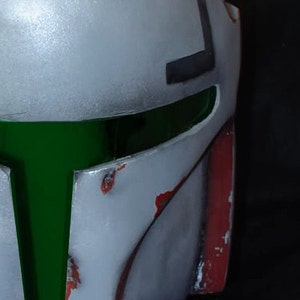 May include: A white and green helmet with red accents and a silver attachment. The helmet has a green visor and a worn, distressed look.