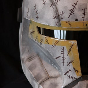 May include: A white and yellow helmet with a visor and brown markings. The helmet has a worn and weathered look, with scratches and scuffs.