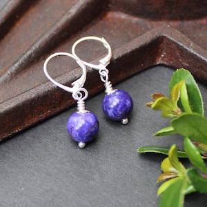 May include: A pair of leverback earrings featuring round, deep purple, mottled stone beads. Each bead is secured with silver-toned wire wrapping that connects to a simple silver-toned metal leverback clasp, creating a dangling design.