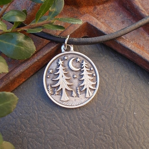 May include: A silver pendant necklace featuring a detailed forest scene with trees, stars, and a crescent moon. The round pendant hangs from a black cord. The design is intricate and nature-themed.