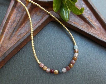 May include: A gold chain necklace with a variety of colored gemstone beads. The beads are in shades of pink, brown, blue, and gold. The necklace is lying on a rustic, brown surface.