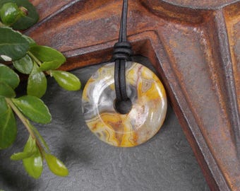 May include: Donut-shaped pendant made of polished stone, featuring a mix of yellow, gray, and white colors. The pendant is strung on a black leather cord. The stone has a natural, marbled appearance, and the pendant is set against a rustic background.