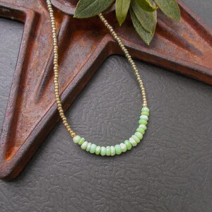 May include: A delicate necklace featuring a gold chain and a strand of small, light green beads. The beads are a soft, pastel green color, and the gold chain adds a touch of elegance. The necklace is displayed on a rustic, brown metal surface.