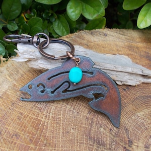 May include: A copper-colored metal fish keychain with a turquoise gemstone charm. The fish is detailed with a cut-out design and has a rustic finish.