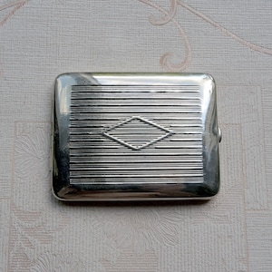 May include: A rectangular silver-toned metal cigarette case featuring a polished finish with horizontal engraved lines across the surface and a centered diamond-shaped outline. The case has rounded corners and a smooth, reflective metallic texture.
