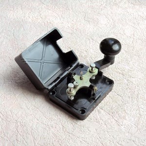 May include: A black telegraph key with a hinged protective cover. The device features a metallic cross-shaped contact arm, silver-colored adjustment screws, and a smooth, round black knob for manual operation, all mounted on a square base.