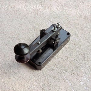 Pode incluir: Um manipulador telegráfico preto com base retangular, braço de alavanca de metal e botão redondo. O dispositivo inclui parafusos ajustáveis, um mecanismo de pivô e orifícios de montagem nos cantos da base de material escuro e polido.