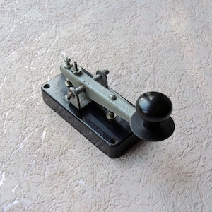 Puede incluir: Una llave telegráfica vintage con una base negra y una palanca plateada. La llave tiene un botón negro. Este dispositivo de comunicación antiguo probablemente está hecho de metal y es un ejemplo clásico de tecnología temprana.