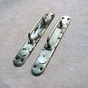 May include: Two vintage metal door handles with a distressed, aged appearance. The handles are silver-toned with peeling paint and rust, featuring a curved bar for gripping. Each handle has a rectangular base with screw holes.