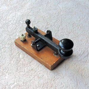Könnte beinhalten: Vintage-Telegrafieschlüssel mit schwarzem Hebel und Knopf, montiert auf einem Holzsockel. Der Schlüssel hat einen schwarzen Metallarm und eine weiße Keramikkomponente. Ein klassisches Kommunikationsgerät.