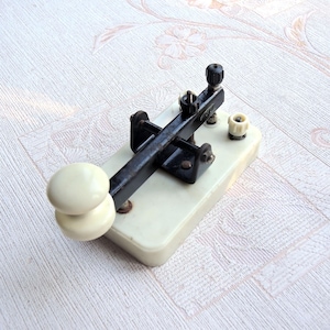 Puede incluir: Clave telegráfica vintage en negro y crema. La llave tiene una palanca y hardware negros, montados sobre una base color crema. Cuenta con dos perillas color crema y una perilla negra en la parte superior.