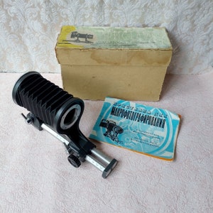 Peut inclure: Un tube d'extension d'appareil photo à soufflets noir avec une base en métal argenté et un rail coulissant. Le tube est fixé à une base en métal argenté avec un bouton pour régler la longueur du tube. Le tube est étiqueté "ПРИСТАВКА ДЛЯ МАКРОФОТОГРАФИРОВАНИЯ".