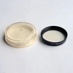 Op de afbeelding: Een doorzichtige plastic lensdop en een zwart lensfilter met wit filtermateriaal. Het lensfilter heeft de tekst "УФ-1× 40,5×0,5". De lensdop is beige en heeft een cirkelvormig ontwerp.