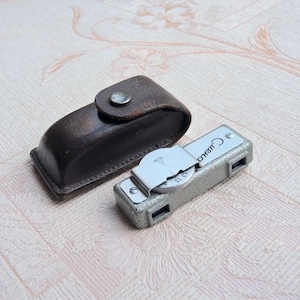 Op de afbeelding: Een vintage lichtmeter met een zilverkleurige behuizing en een wijzerplaat, naast een bruine leren etui met een drukknoopsluiting. De meter heeft het woord "Смена" erop.