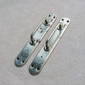 Könnte beinhalten: Zwei antike silberfarbene Türgriffe. Jeder Griff hat eine lange, rechteckige Basis mit einem gebogenen Griff und einem kreisförmigen Detail. Das Metall weist Gebrauchsspuren auf, mit einigen Bereichen mit Farbverlust, was ihnen ein rustikales Aussehen verleiht.