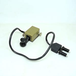 May include: An antique telegraph key with a black knob and a khaki-colored metal body. A black wire connects the key to a black electrical plug. The key has two silver-colored screws on the sides.