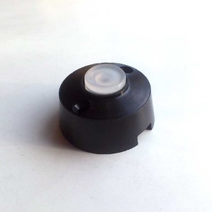 May include: A black plastic base with a white, round, translucent button on top. The base has a small hole on the side.