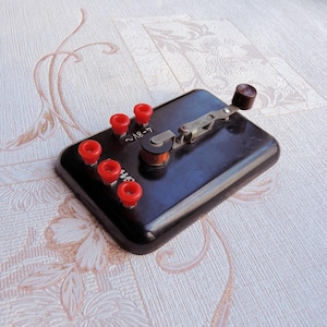 Può includere: Un dispositivo elettrico vintage nero con una manopola rossa e cinque terminali rossi. Il dispositivo ha una leva in metallo e una piccola bobina. Il testo "L-8V-2" è stampato sul dispositivo.