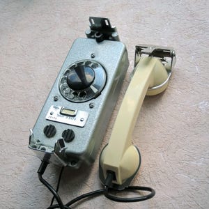 Puede incluir: Teléfono de pared plateado vintage con un dial giratorio y un auricular crema. El teléfono tiene un cuerpo rectangular con un dial, botones y una pequeña pantalla. El auricular está conectado por un cable negro.