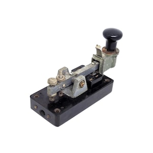 Può includere: Chiave telegrafica vintage con base nera e leva color argento. La chiave ha un pomello nero e vari componenti metallici, tra cui viti e molle. Il dispositivo è progettato per l'invio del codice Morse, su sfondo bianco.