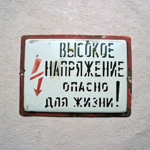 May include: A rectangular metal sign with a white background and a red border. The sign features black Cyrillic text and a red lightning bolt symbol, indicating a high voltage warning. The sign is aged and shows signs of wear.