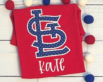 May include: Red sleeveless shirt with a blue and white polka dot St. Louis Cardinals logo and the name "Katie" embroidered in white.