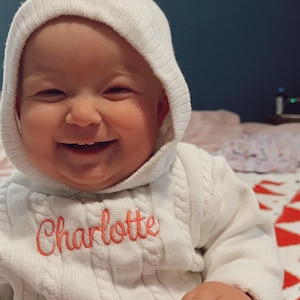 May include: A baby girl wearing a white knitted hooded sweater with the name "Charlotte" embroidered in orange on the front. The baby is smiling and sitting on a bed.