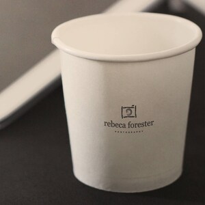 May include: White paper cup with a black and white logo that says "rebecca forester photography" and a camera icon.