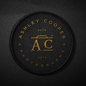 Puede incluir: Un parche circular negro con texto y dise&ntilde;o bordados en oro. El texto dice "ASHLEY COOPER PHOTOGRAPHY" alrededor del borde, con "ESTD 2013" y "AC" con un icono de c&aacute;mara en el centro. El parche tiene una apariencia texturizada.