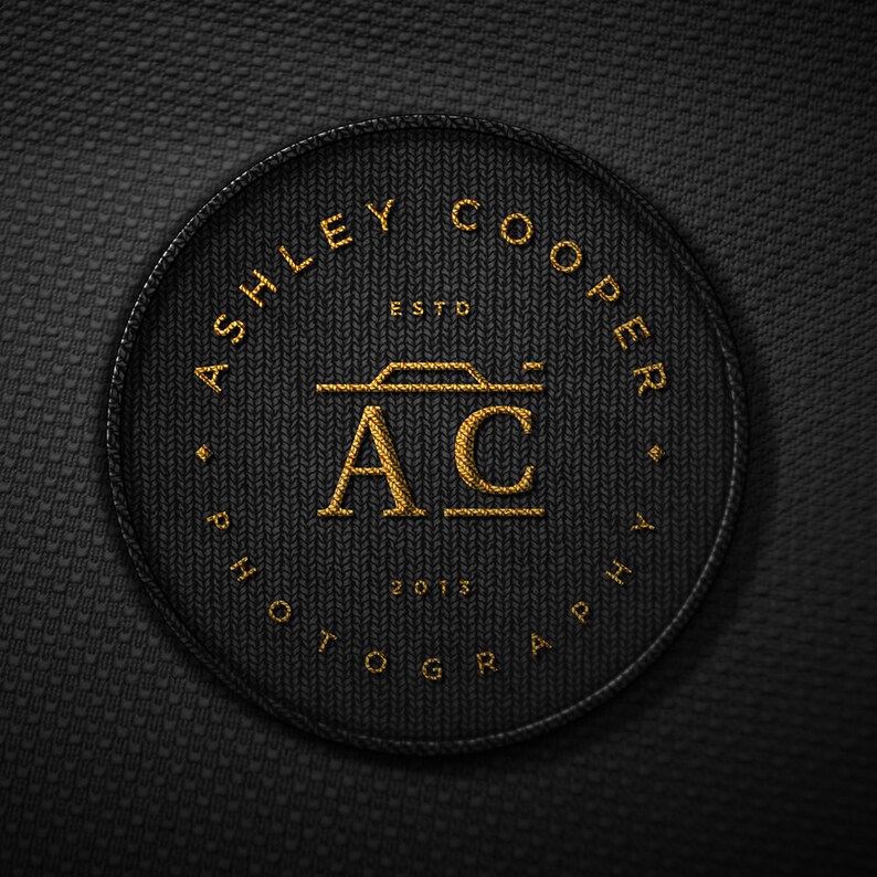 Puede incluir: Un parche circular negro con texto y dise&ntilde;o bordados en oro. El texto dice "ASHLEY COOPER PHOTOGRAPHY" alrededor del borde, con "ESTD 2013" y "AC" con un icono de c&aacute;mara en el centro. El parche tiene una apariencia texturizada.