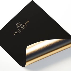 Puede incluir: Tarjeta negra con interior de l&aacute;mina dorada, con el texto dorado "AC ASHLEY COOPER PHOTOGRAPHY". La tarjeta est&aacute; ligeramente curvada, revelando la brillante l&aacute;mina dorada debajo.