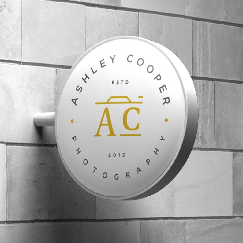 Puede incluir: Un letrero circular blanco con el texto "ASHLEY COOPER PHOTOGRAPHY" dispuesto en c&iacute;rculo. El centro presenta "AC" en letras doradas, un icono de c&aacute;mara y "ESTD 2013". El letrero est&aacute; montado en una pared de ladrillos grises.