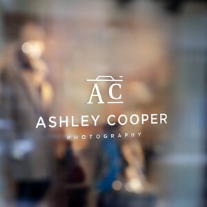 Puede incluir: Logotipo blanco sobre una superficie de vidrio. El logotipo incluye las iniciales "AC" con un icono de c&aacute;mara encima y el nombre "ASHLEY COOPER PHOTOGRAPHY" debajo. El fondo est&aacute; borroso, sugiriendo un entorno exterior.