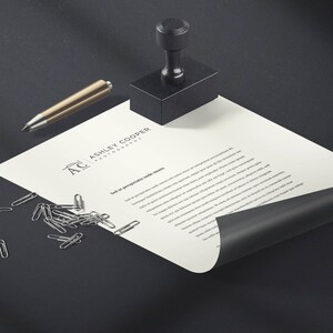 Puede incluir: Un juego de papeler&iacute;a con un sello negro, un bol&iacute;grafo dorado y clips plateados sobre una superficie negra. Tambi&eacute;n hay una hoja de papel con el texto "ASHLEY COOPER PHOTOGRAPHY". El papel est&aacute; ligeramente curvado.