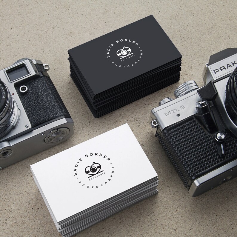 May include: Stacks of black and white business cards with the text "Sadie Border Photography ESTD-2017" and a camera logo. Two vintage cameras are positioned on either side of the cards.