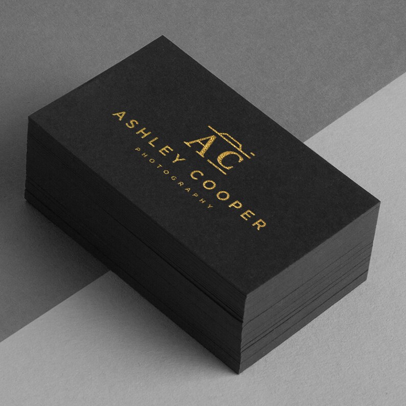 Puede incluir: Una pila de tarjetas de visita negras con letras doradas. El texto dice "ASHLEY COOPER PHOTOGRAPHY" con las iniciales "AC" y un icono de c&aacute;mara. Las tarjetas est&aacute;n apiladas sobre una superficie gris.