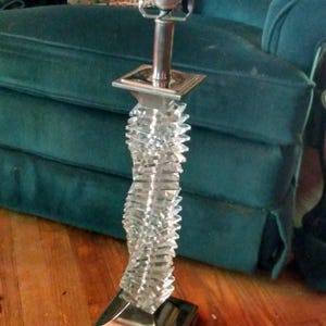 May include: A clear, decorative table lamp with a silver-colored metal base and accents. The lamp features a twisted, textured glass column and a silver lamp holder. The lamp is approximately 24 inches tall.