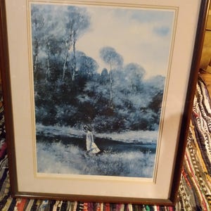 May include: Framed landscape art print featuring a serene scene with a figure in a white dress near a body of water. The artwork is dominated by shades of blue and white, with trees and a cloudy sky. The frame is a dark brown color.