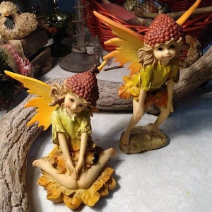 May include: Two fairy figurines. One is seated on a yellow flower, the other is standing. Both have brown acorn-shaped hats, yellow and orange wings, and green and orange clothing. The figurines are detailed with delicate facial features and are posed in a whimsical manner.