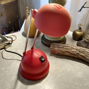 May include: A red desk lamp with a rounded shade and base. The lamp has a silver adjustable arm and a black on/off switch. The shade has three slits on top for ventilation. The lamp is on a table.