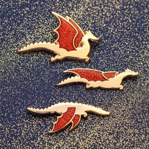 May include: Three gold-colored dragon pins with red glitter wings and accents are displayed on a dark blue background with sparkling glitter. The dragons are in a flying pose, with detailed features.