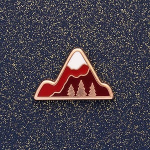 May include: A mountain-shaped enamel pin with a red and white design. The pin features a mountain range with three green trees at the base and a gold-coloured border. The background is a dark blue with gold flecks.