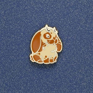May include: A gold-toned metal enamel pin shaped like a lop-eared rabbit. The rabbit features a cream-coloured body with patches of reddish-brown fur, long drooping ears, and a small tail. The design includes fine gold outlines defining the features.
