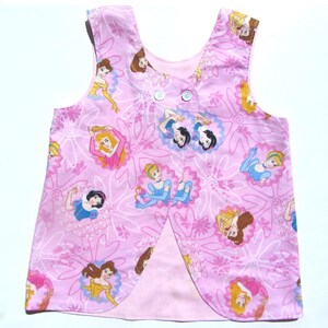 May include: Pink sleeveless top with a princess pattern. The fabric features various princess illustrations within floral frames, set against a pink background. The top has a button closure and a split front design.