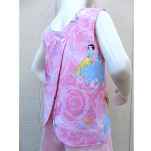 May include: Pink sleeveless top with a princess print and a rose pattern. The top has a button closure and is designed for children. The fabric is a light pink color with a repeating pattern of roses and butterflies.