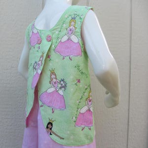 May include: A light green vest with a princess pattern. The vest features a wrap-around design with pink buttons and a pink and white princess print. The princess print includes crowns, flowers, and the word "princess".