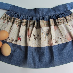 May include: A blue denim apron with a brown and white checkered trim and a cream fabric panel featuring a farm animal print. Three brown eggs are in front of the apron.