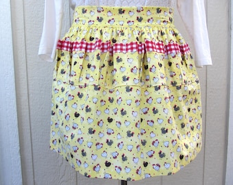 May include: Yellow apron with a repeating pattern of chickens in black, white, and red. The apron has a red and white checkered band and a ruffled design. The apron is worn over a white long-sleeved shirt.