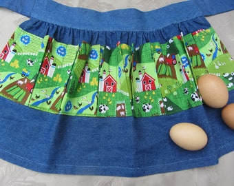 May include: A blue denim apron with a green and white farm-themed print. The apron has a gathered skirt and a tie at the waist. Three brown eggs are sitting on the apron.