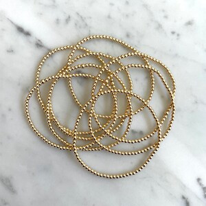 May include: A stack of gold beaded bracelets. The bracelets are made of small, round beads that are strung together on a thin wire. The bracelets are all the same size and are stacked on top of each other.